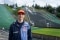 The photo shows Karl Geiger smiling at the camera. Ski jumps can be seen in the background.